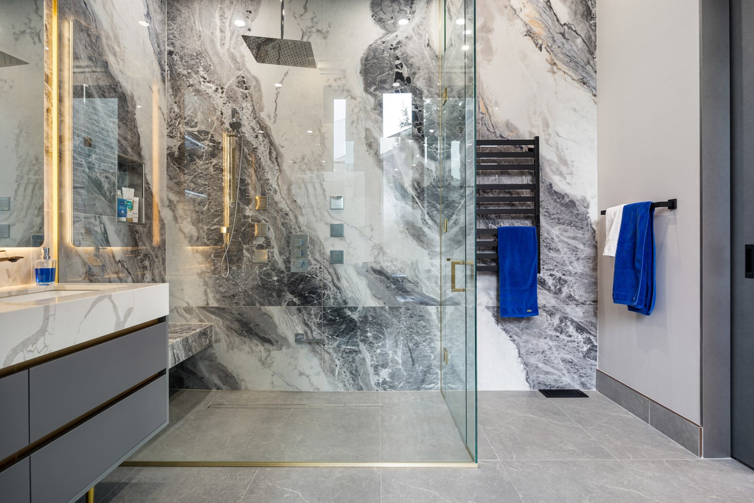 Primary ensuite shower with full-slab book-matched marble walls, ceiling rain head, gold body jets and frameless glass enclosure