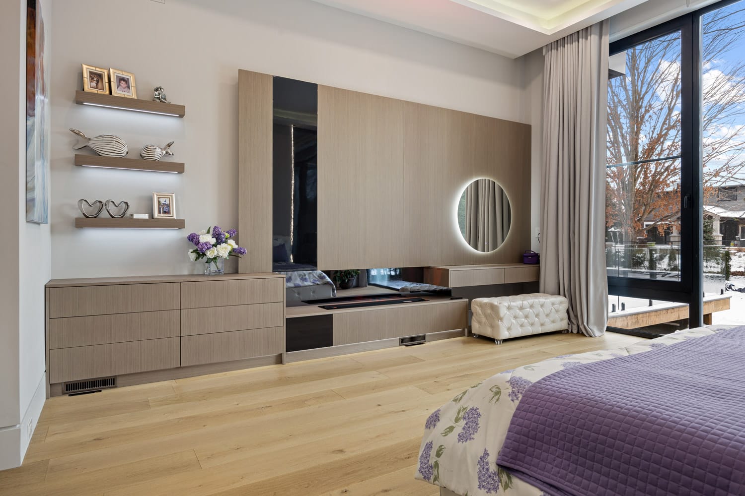 Primary bedroom millwork wall with built-in oak cabinetry, round backlit mirror, floating drawers and private deck access